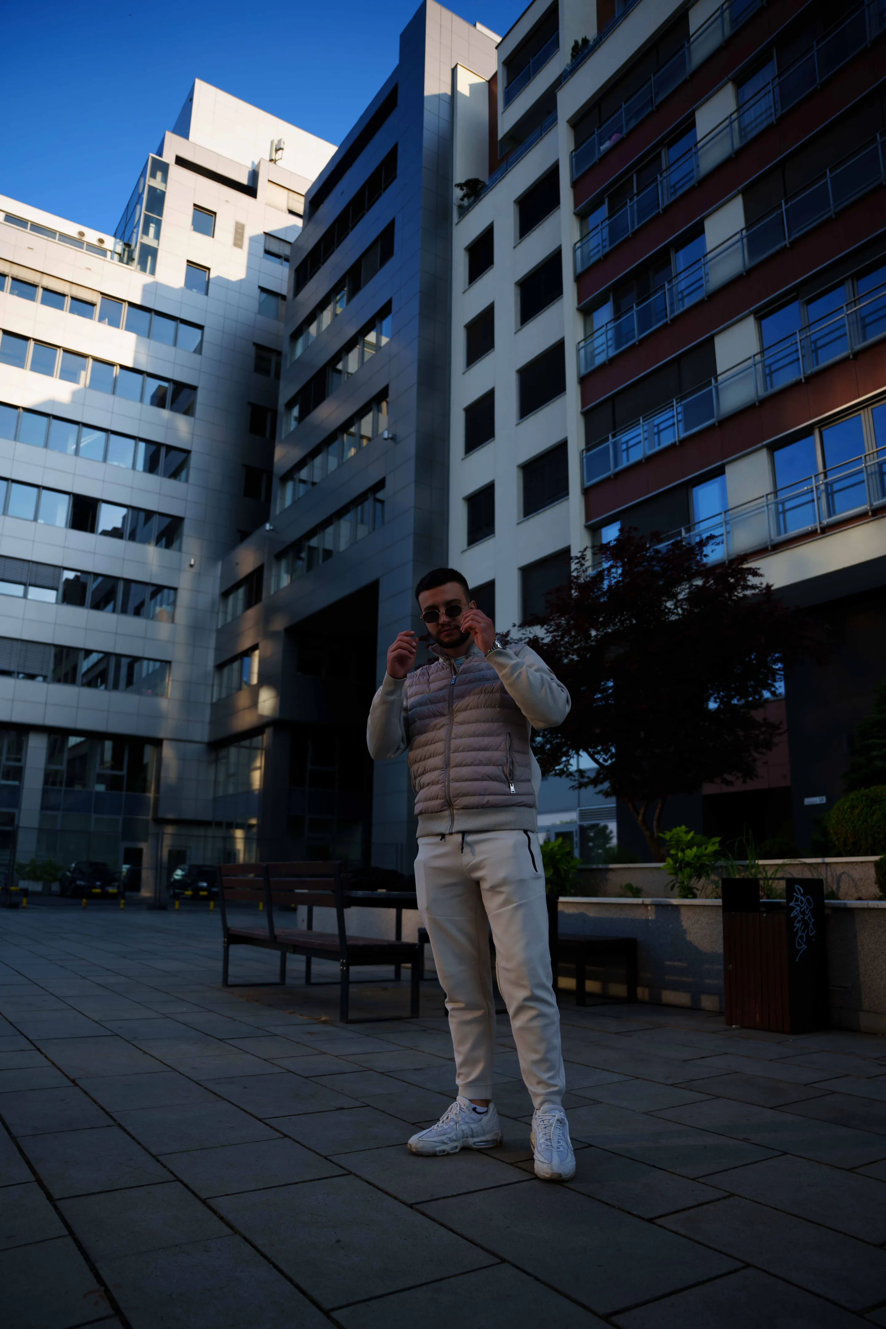 Full length in a beige puffer and cream joggers, adjusting sunglasses in the courtyard of the glass block