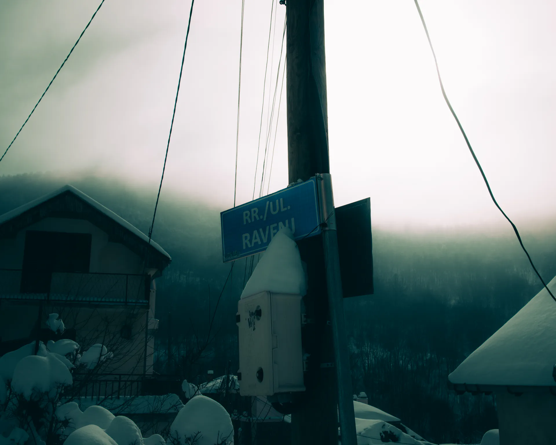 Blue street sign — RR. / UL. RAVENI — snow-capped on its pole, the village behind fog