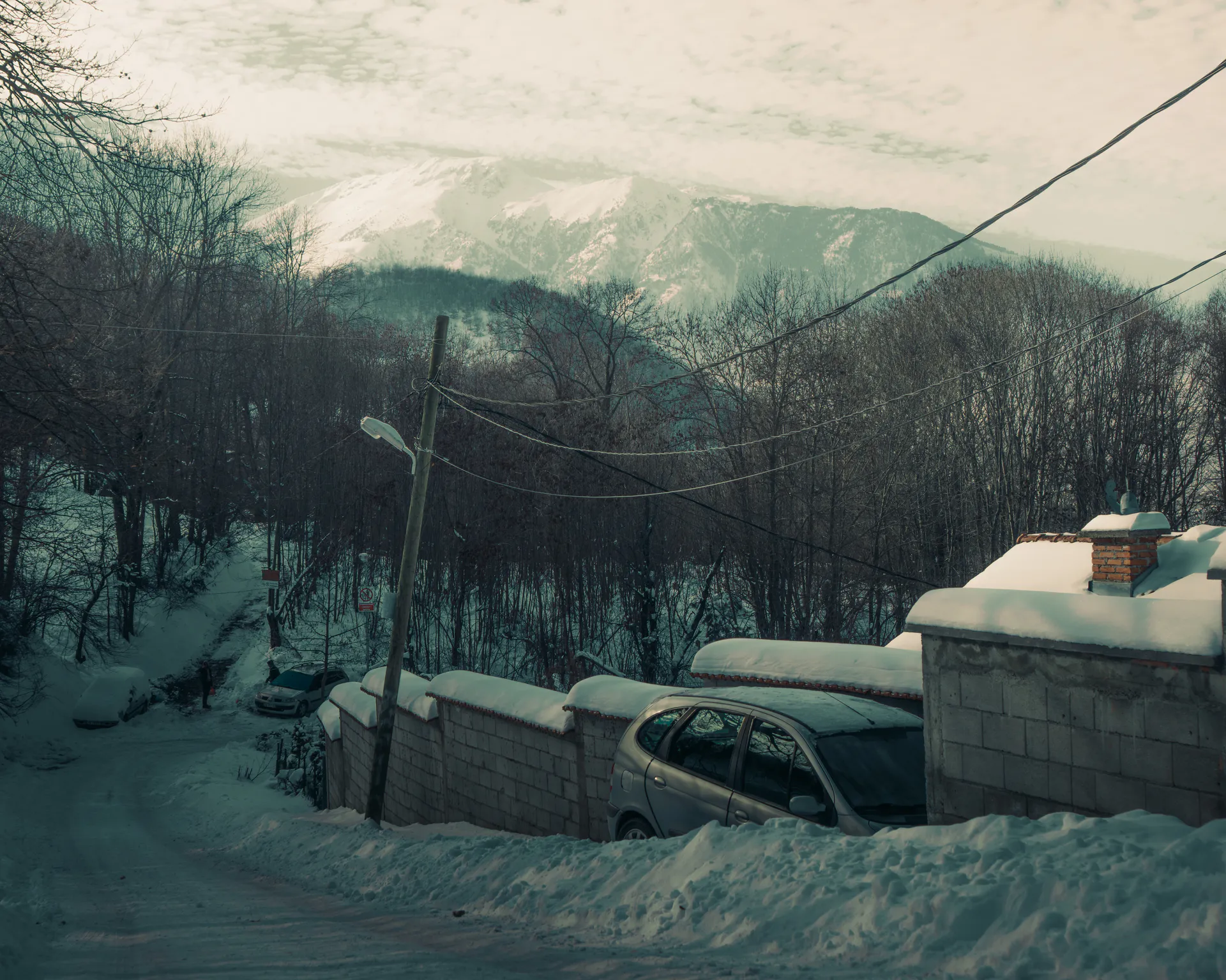 A parked car beside the snow-topped block wall, chimney and forested slopes above