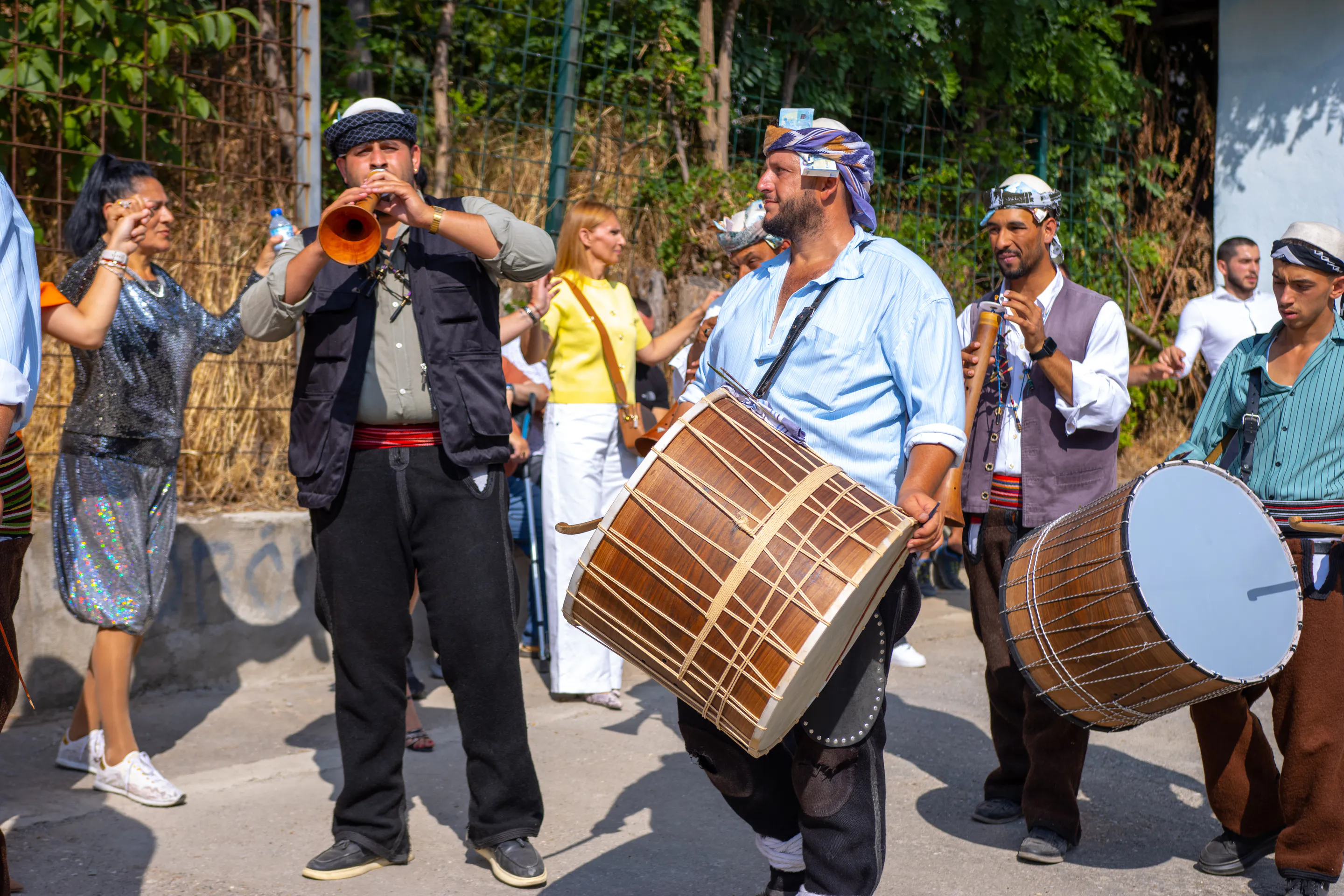 Procession musicians on the village road — two drummers with slung tapan drums and a zurna player leading the guests downhill