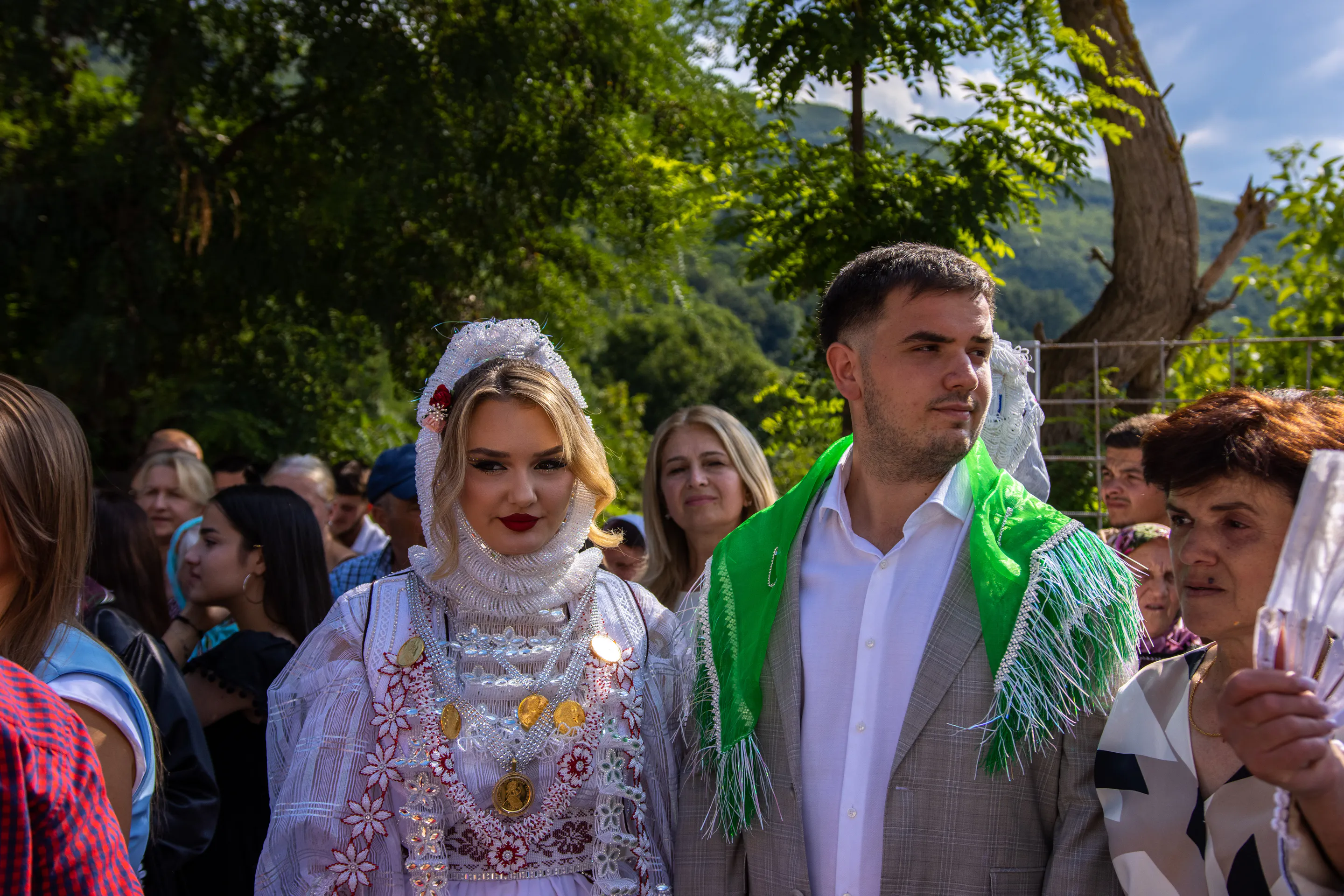The bride-to-be in white beaded traditional dress and gold coins beside her fiancé, guests gathered under the trees