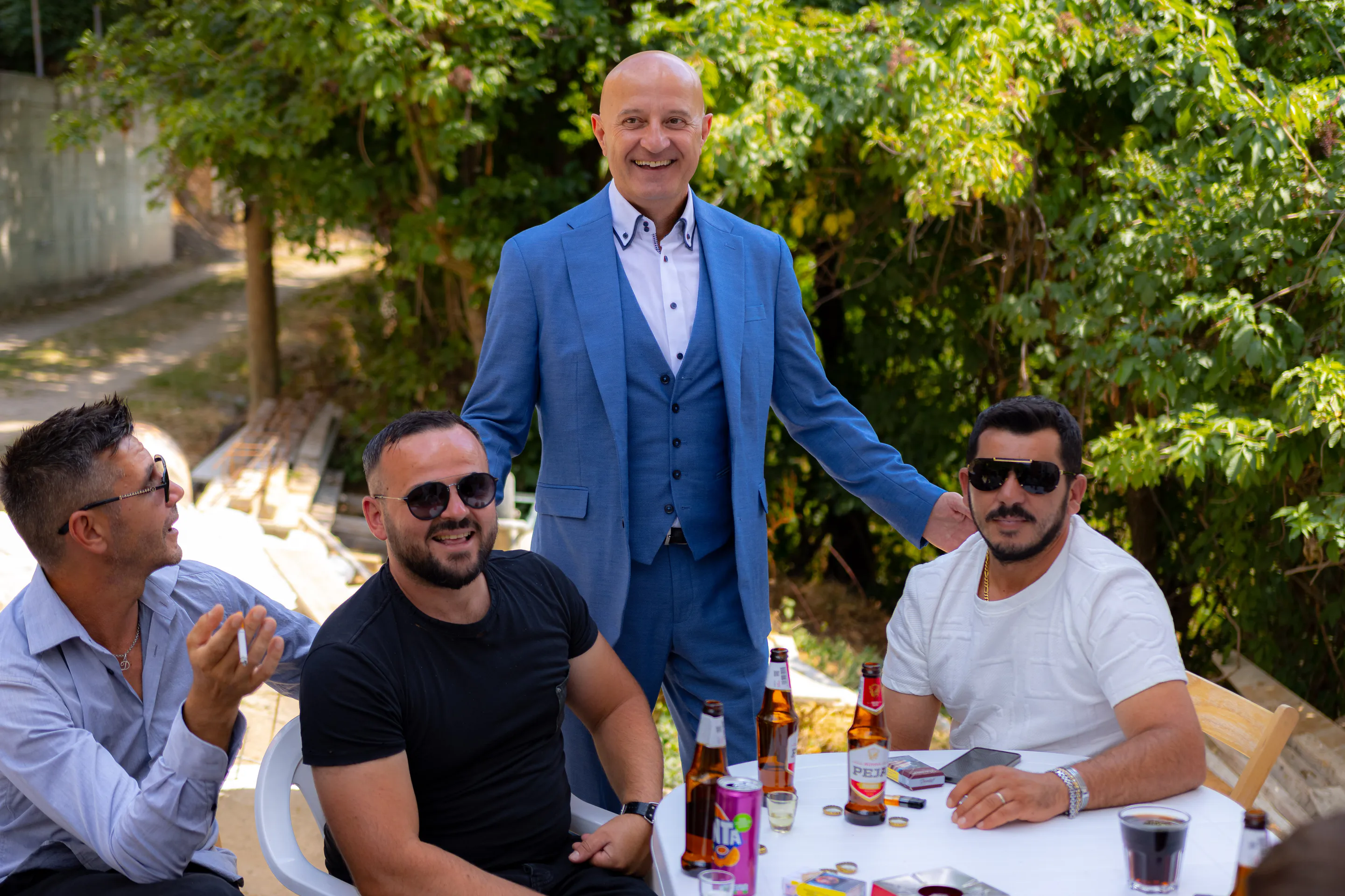 Guests at the garden table, one standing mid-toast in a bright blue suit