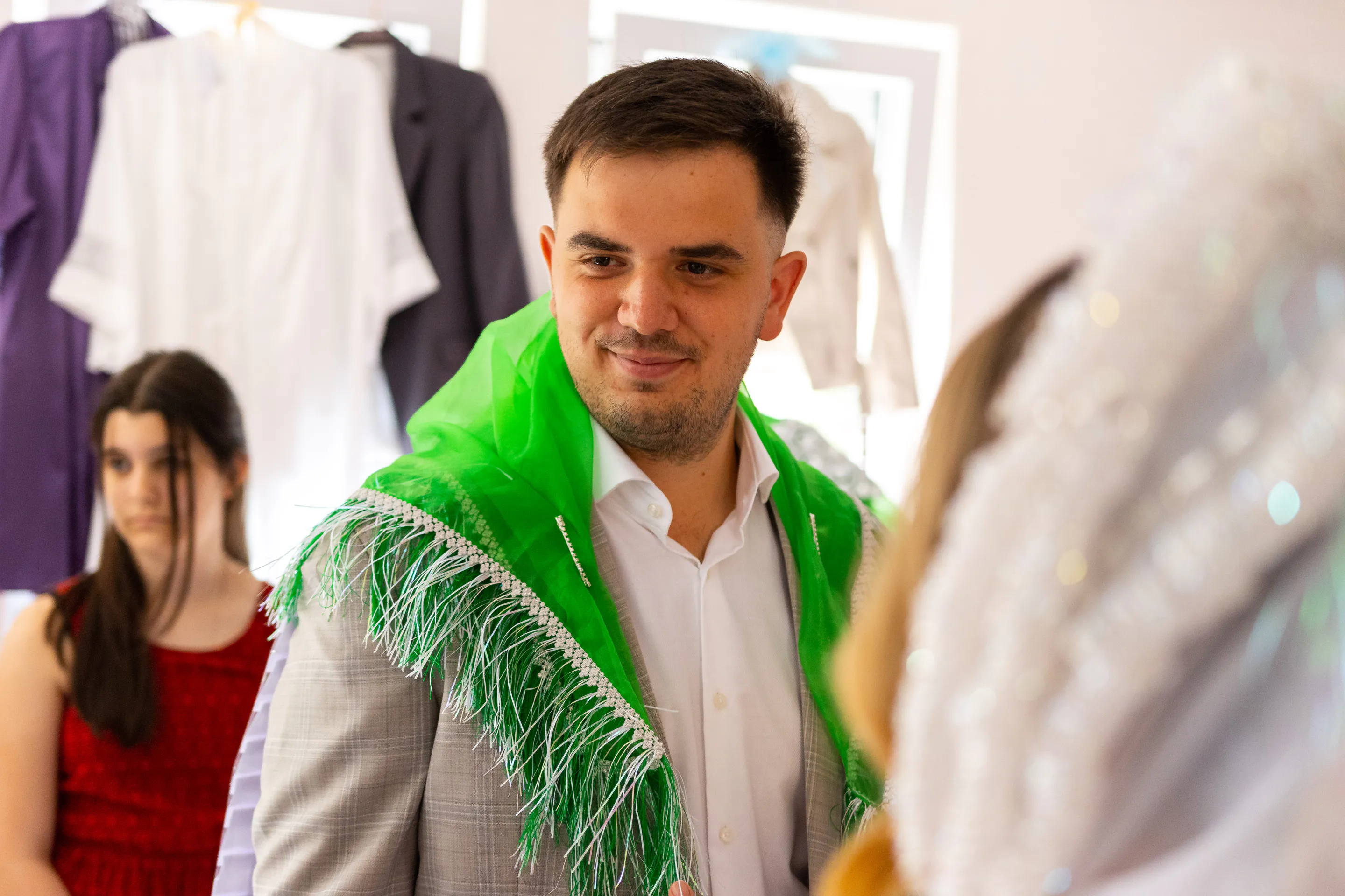The groom-to-be in a pale suit with the green fringed ceremonial shawl, in the dressing room between hanging garments