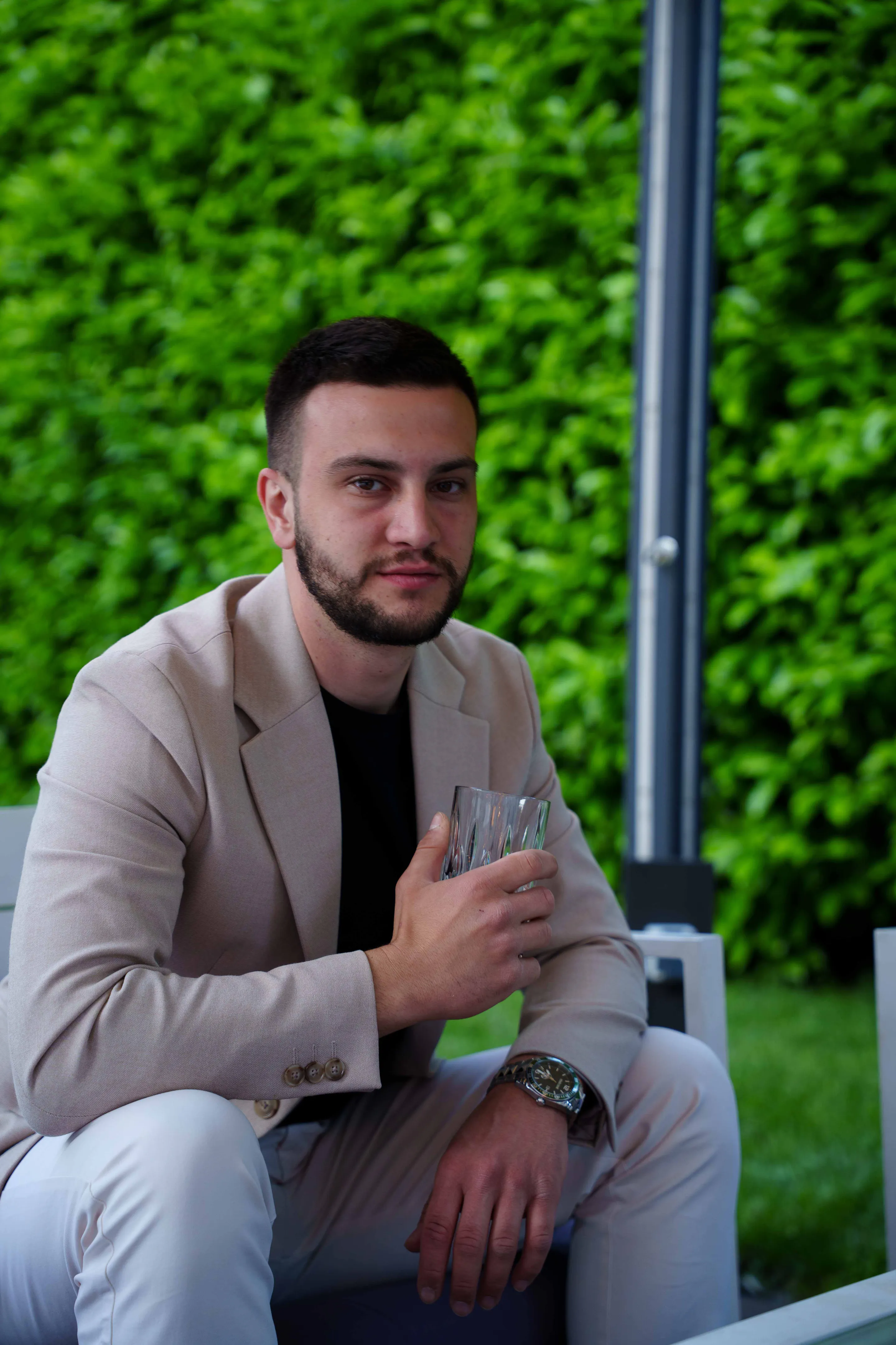 Seated in a cream blazer with a glass, the hedge wall behind