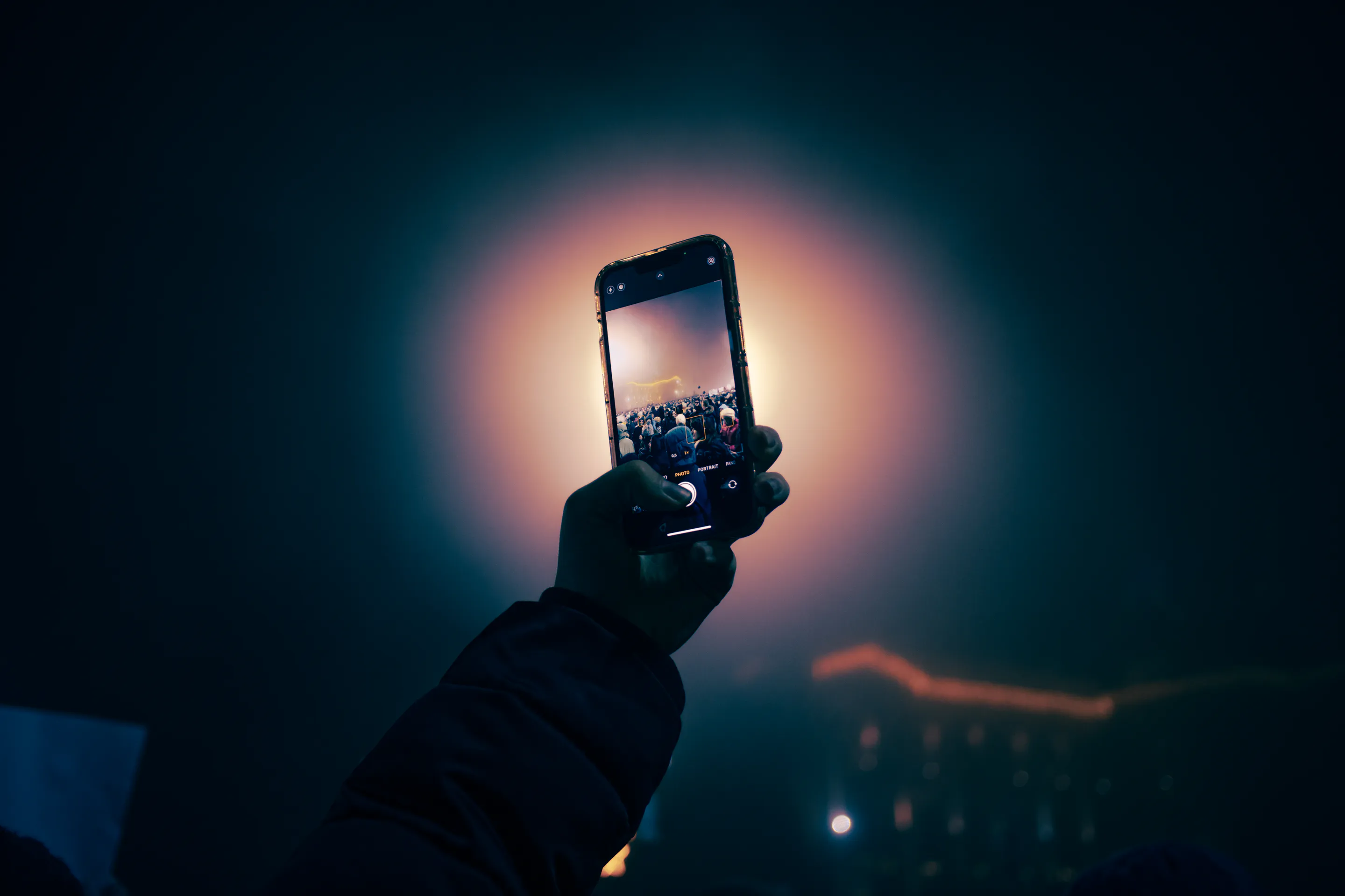 A hand holds a phone up over the crowd, its screen lit against the orange glow