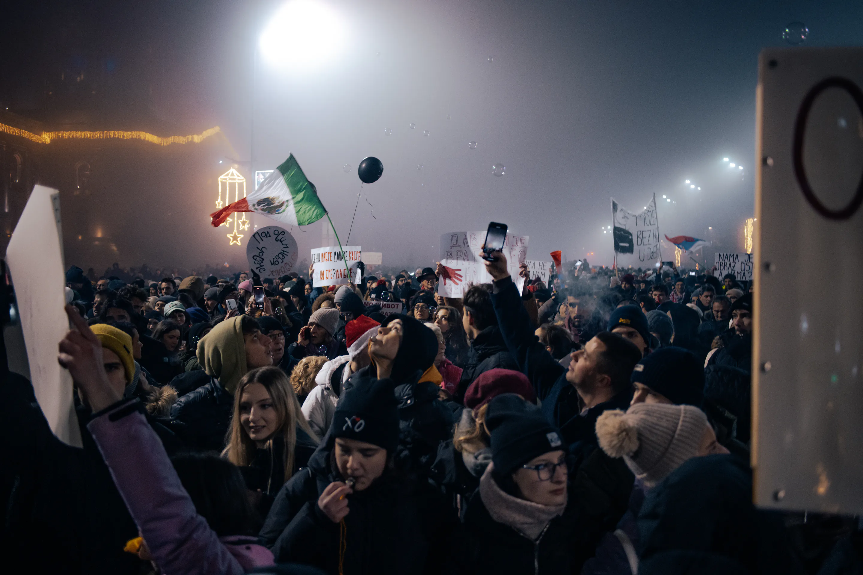 Signs and a flag above the packed street, soap bubbles drifting through the light