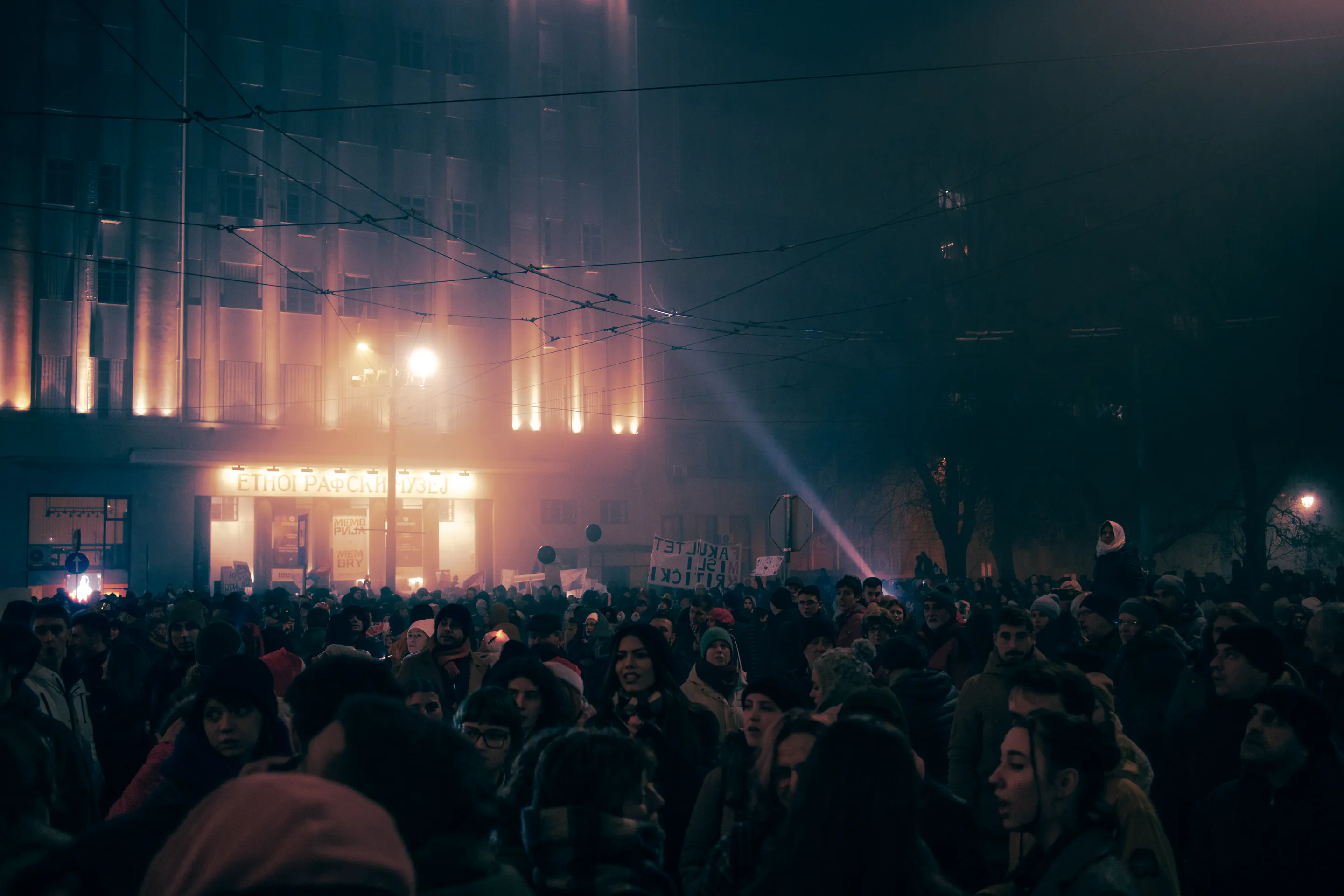 The crowd filling the street before the floodlit Ethnographic Museum, one beam cutting the fog