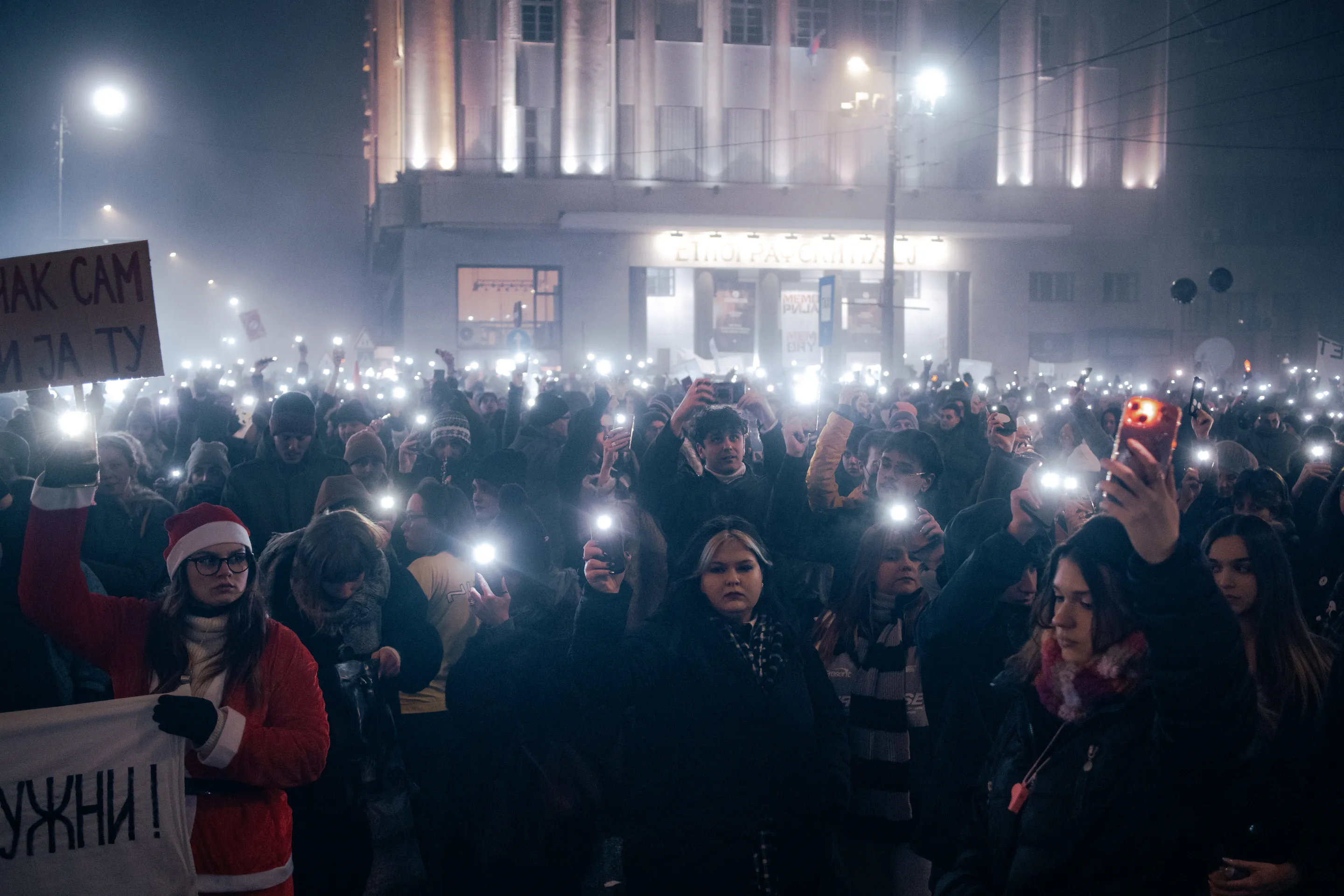 Phone flashlights held overhead; a woman in a Santa suit fronts the crowd with a sign