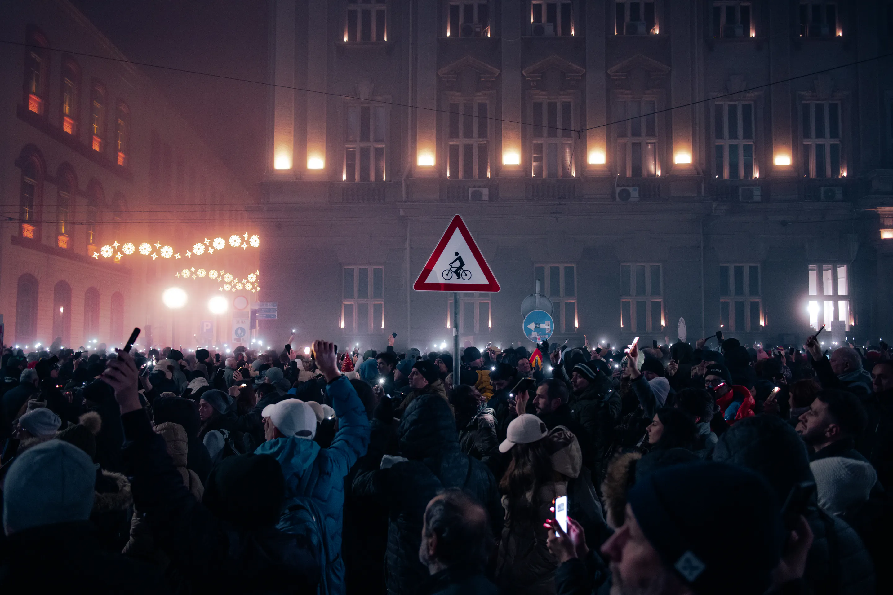 The crowd beneath a lit bicycle-crossing sign, amber windows burning through the fog
