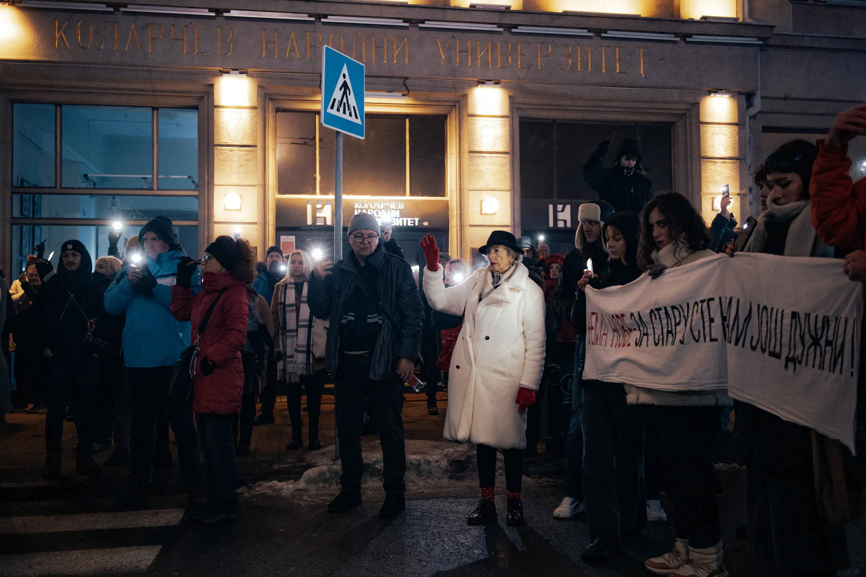 An elderly woman in a white coat raises her hand amid the banner line