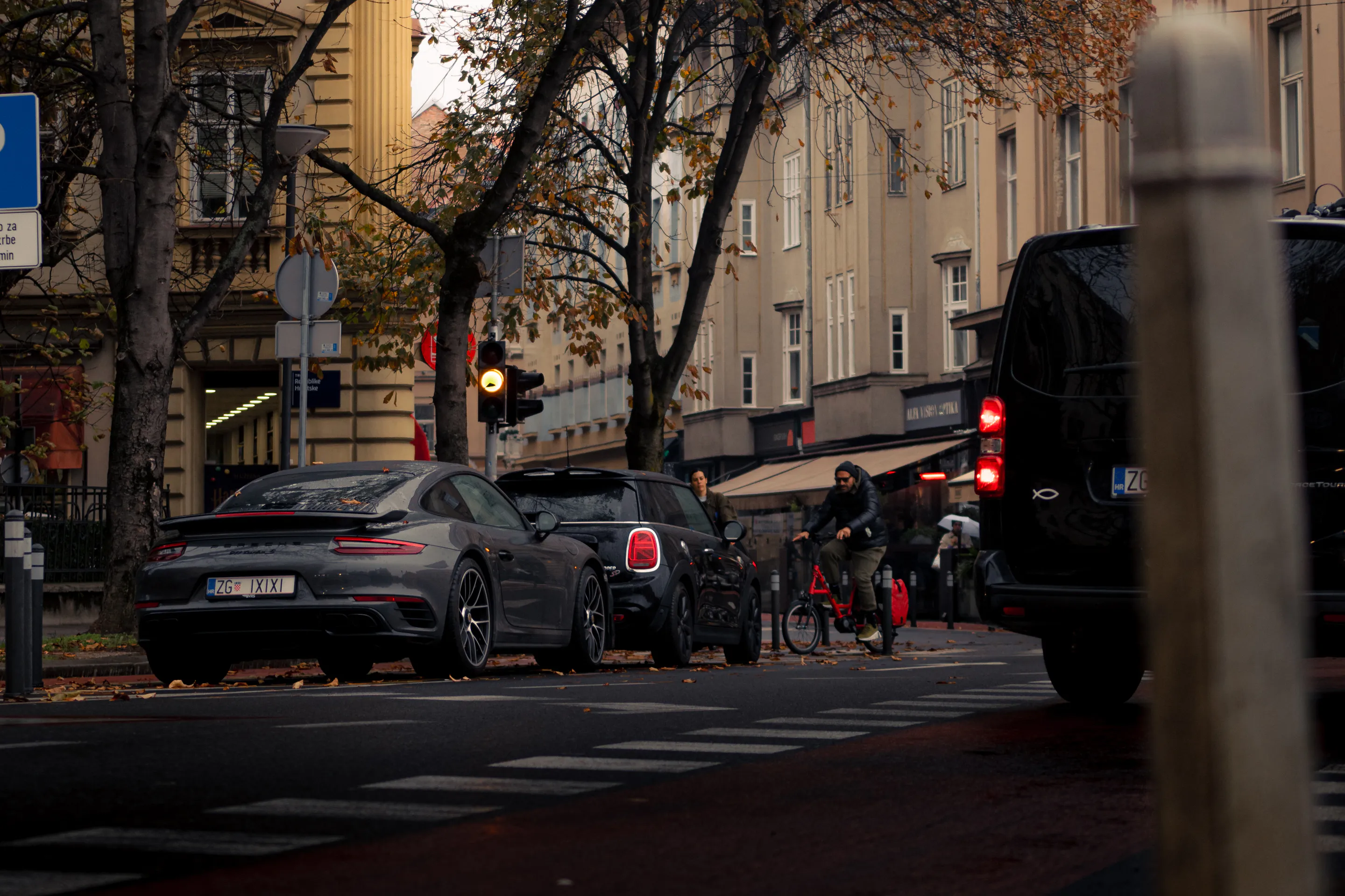 Grey 911 and a cyclist with red panniers held at the crosswalk, autumn trees over pastel facades