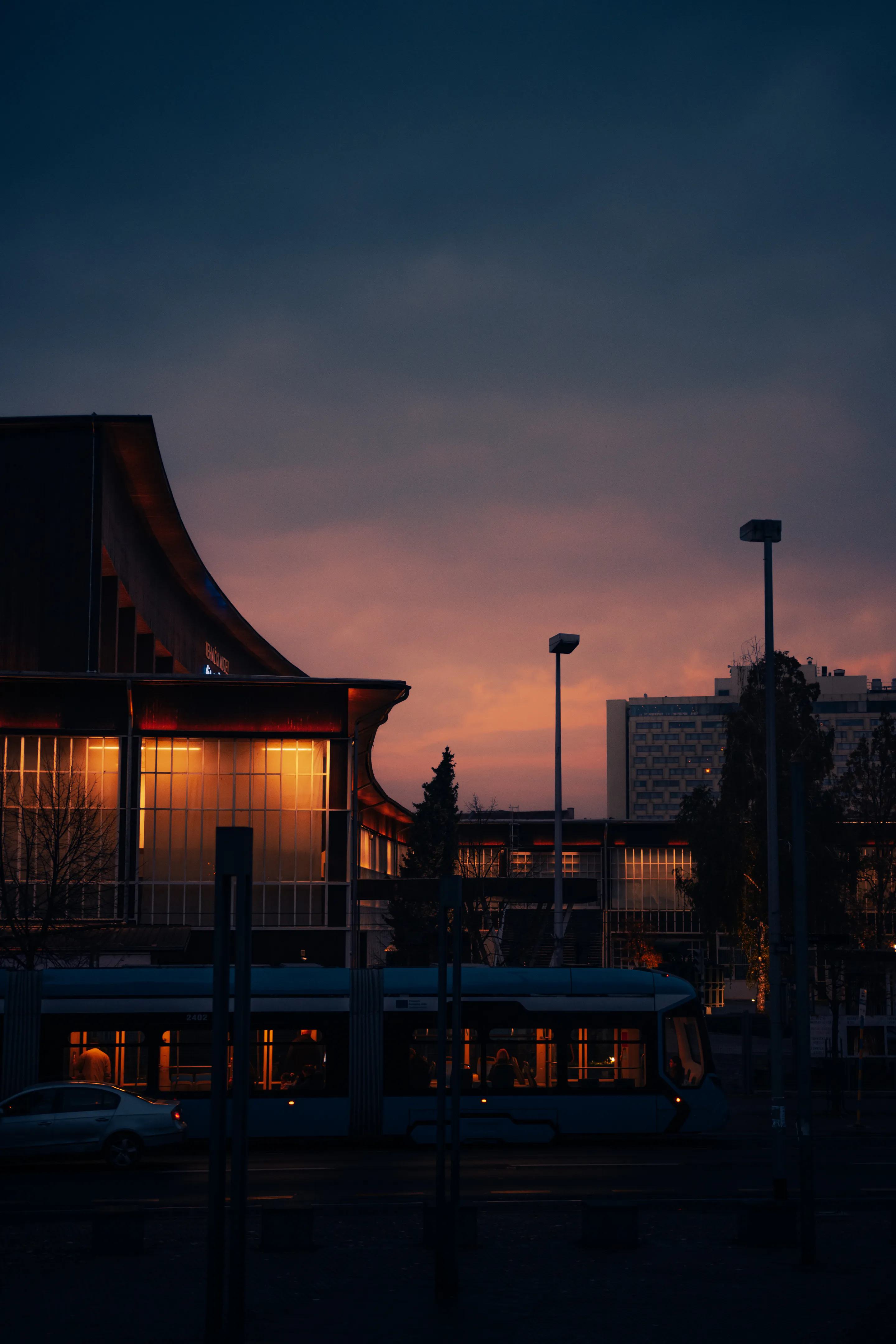 Curved modernist block and a passing tram silhouetted against the orange-to-navy sunset