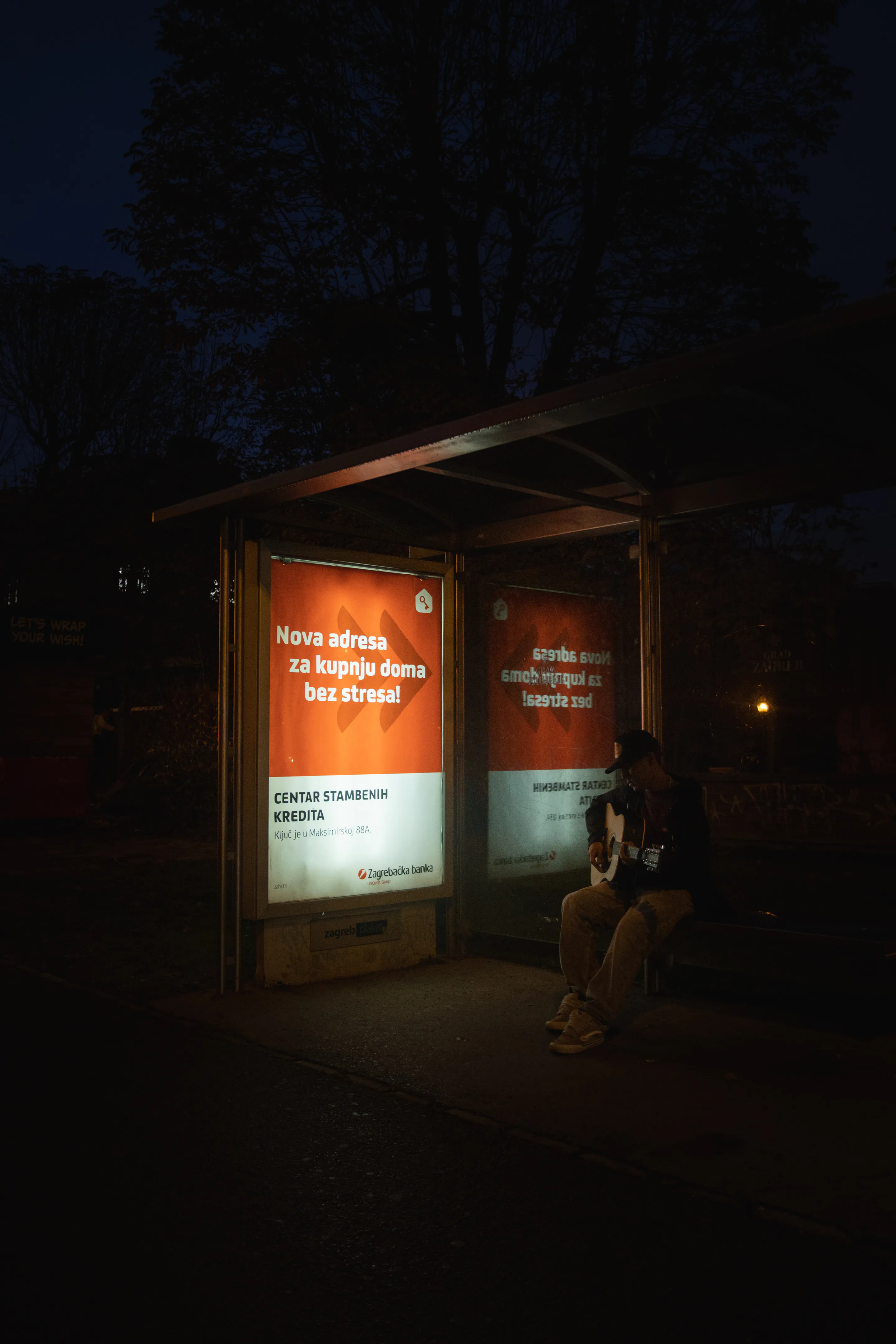 A guitarist alone in the lit bus shelter beside the glowing ad panel